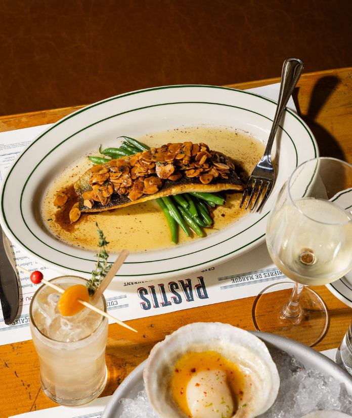 A plated fish fillet topped with toasted almonds and served with green beans in a butter sauce, accompanied by a cocktail, white wine, and a baked egg dish on a restaurant table.