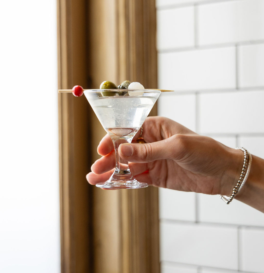 Hand holding a martini glass with olives and a toothpick, white subway tiled wall and brass pole in background.