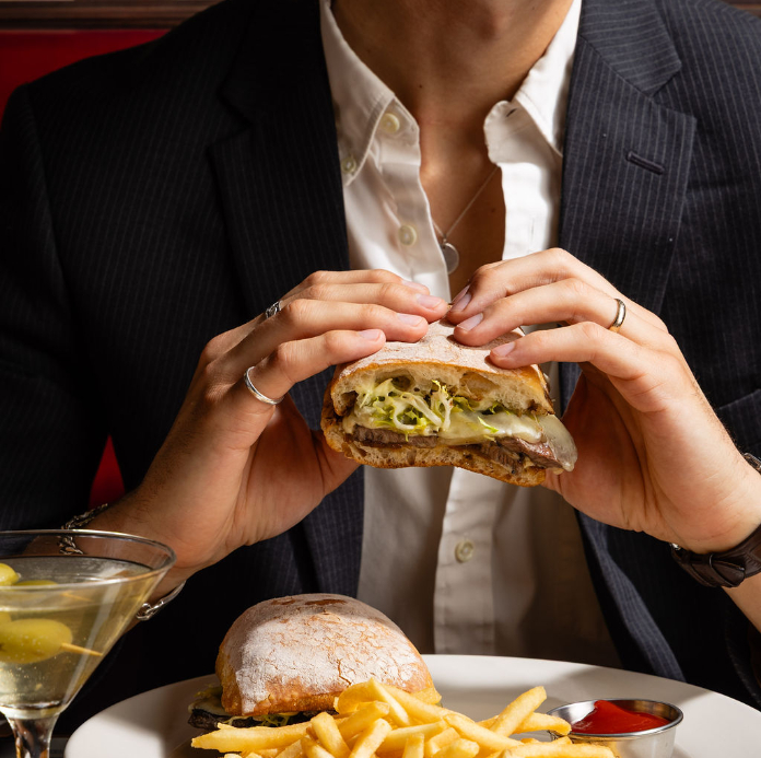 A person in a blazer holding a burger with fries and a martini on the table.