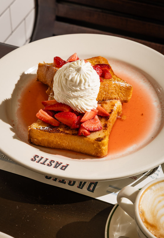 French toast topped with whipped cream and strawberries in strawberry syrup on a white plate with coffee cup beside it