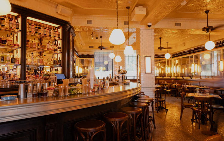 Warmly lit bar with wooden stools, mirrored shelves, and large windows, creating a cozy interior setting.